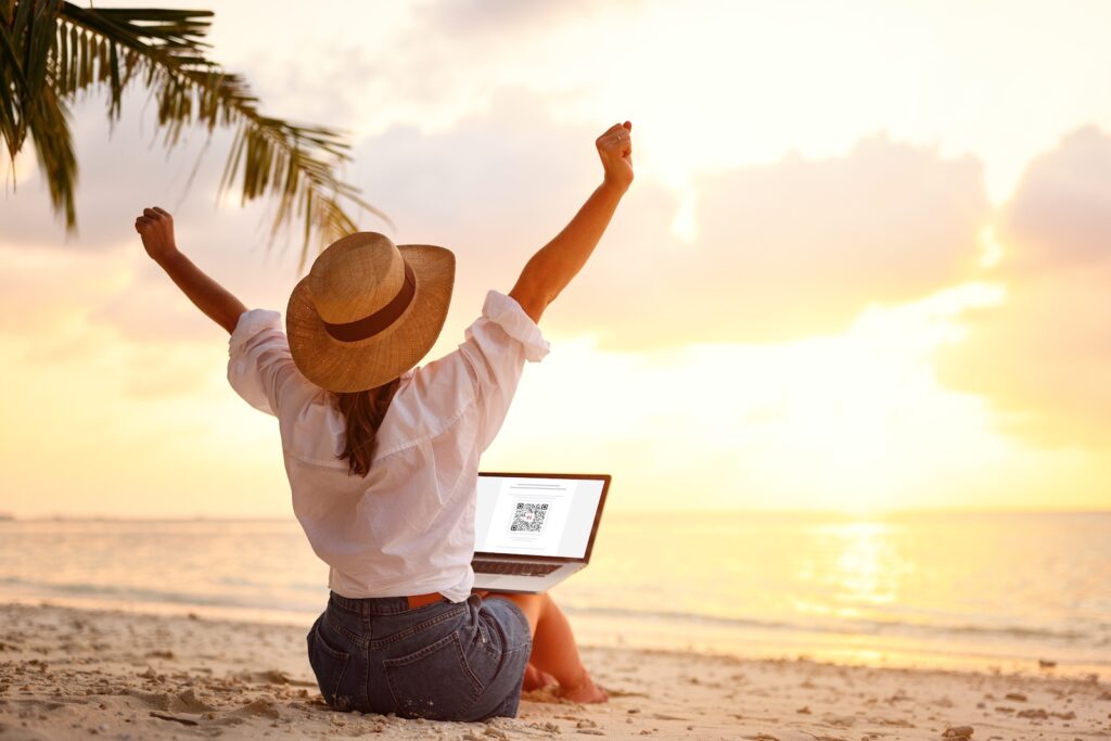 Femme assise sur une plage tropicale au Costa Rica, bras levés de joie devant un coucher de soleil, travaillant sur un ordinateur portable avec un QR code à l’écran — symbole de liberté numérique et de télétravail sous les tropiques.